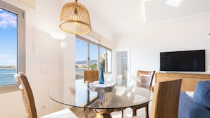 A modern living room with a glass-top dining table, wooden chairs, a blue sofa, a TV, and a view of the sea.