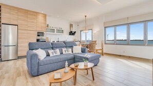 A modern living room with a grey sectional sofa, wooden flooring, and a large window offering a view of the sea.