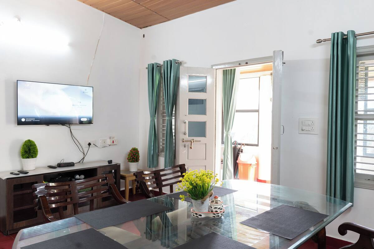 A dining area with a glass table, chairs, a TV mounted on the wall, and a door leading to another room.