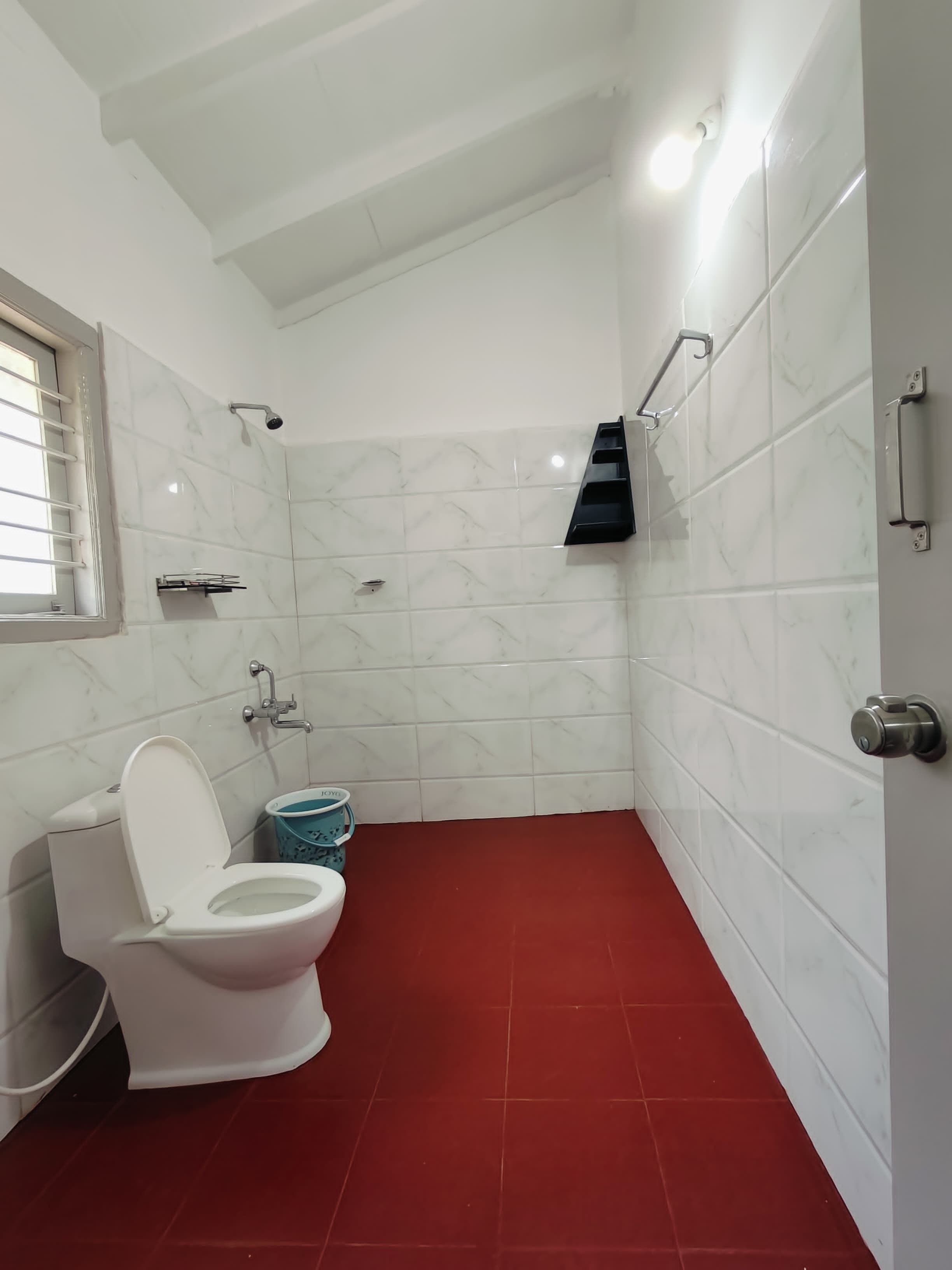 Une salle de bain avec des carreaux de marbre blanc, un sol rouge, des toilettes blanches, une douche et une fenêtre.