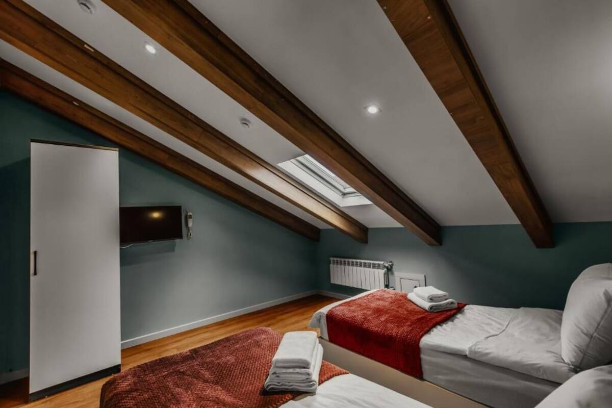 Une chambre avec un plafond en pente, des poutres en bois, un puits de lumière, un lit, un radiateur et une armoire.