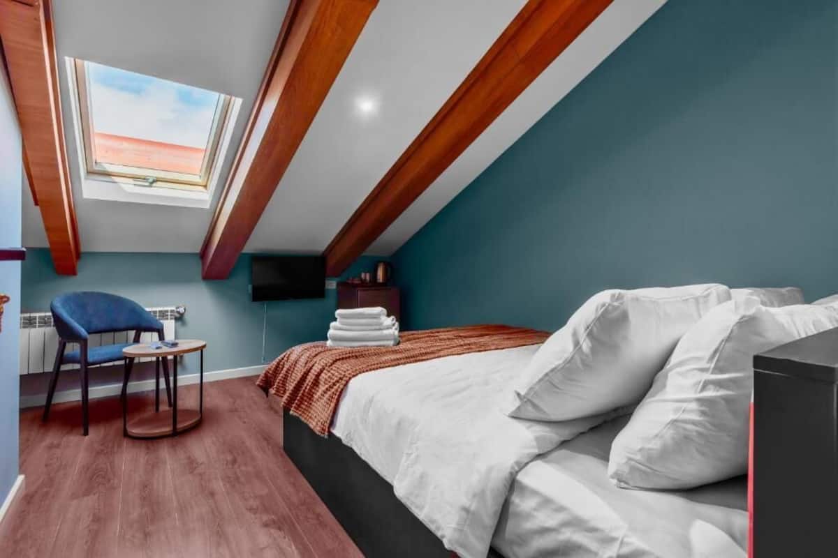 Une chambre avec un puits de lumière, un lit recouvert de draps blancs, une chaise bleue et une petite table.