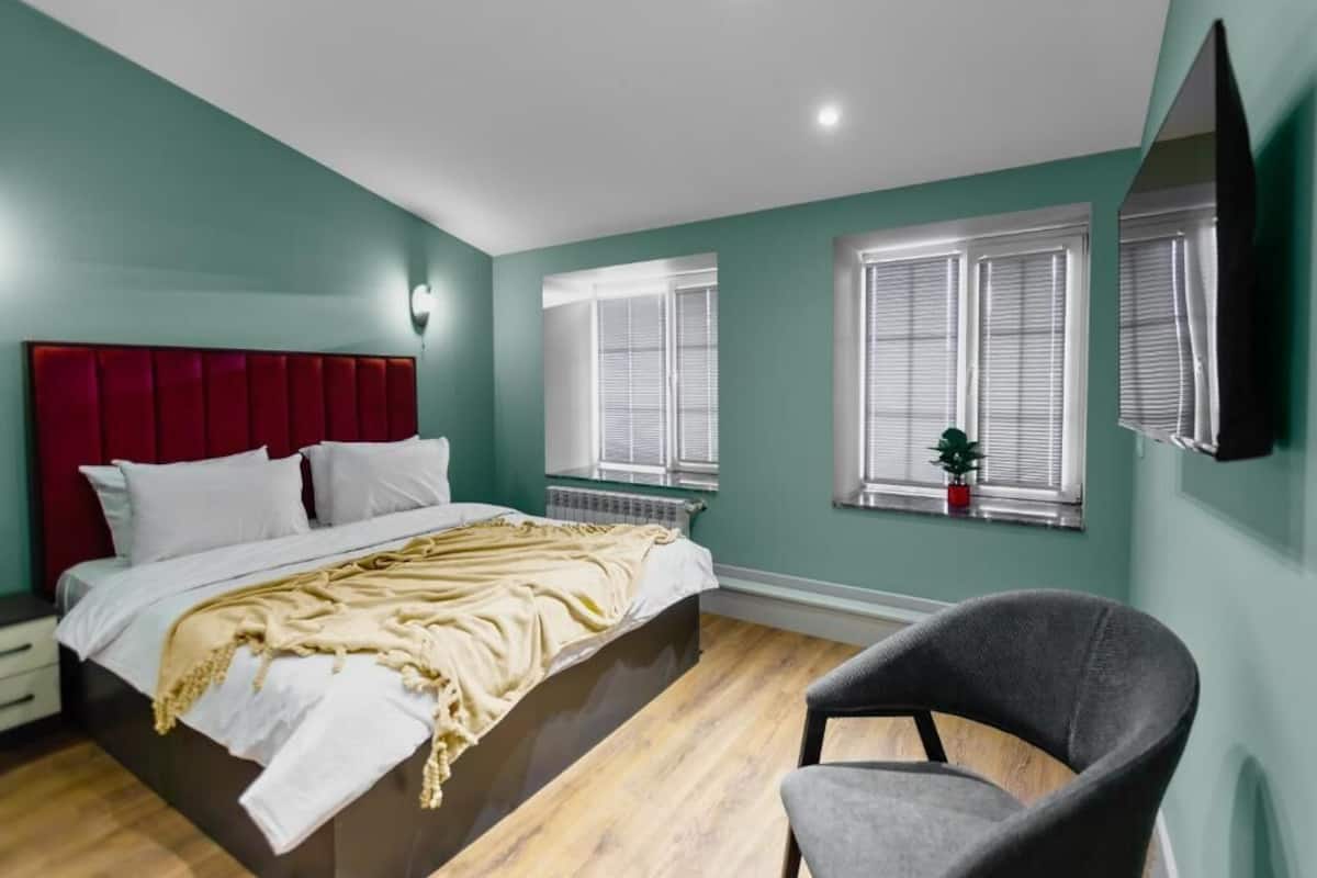 Une chambre à coucher avec une tête de lit rouge, un lit recouvert de draps blancs, un fauteuil gris et une fenêtre avec des stores.