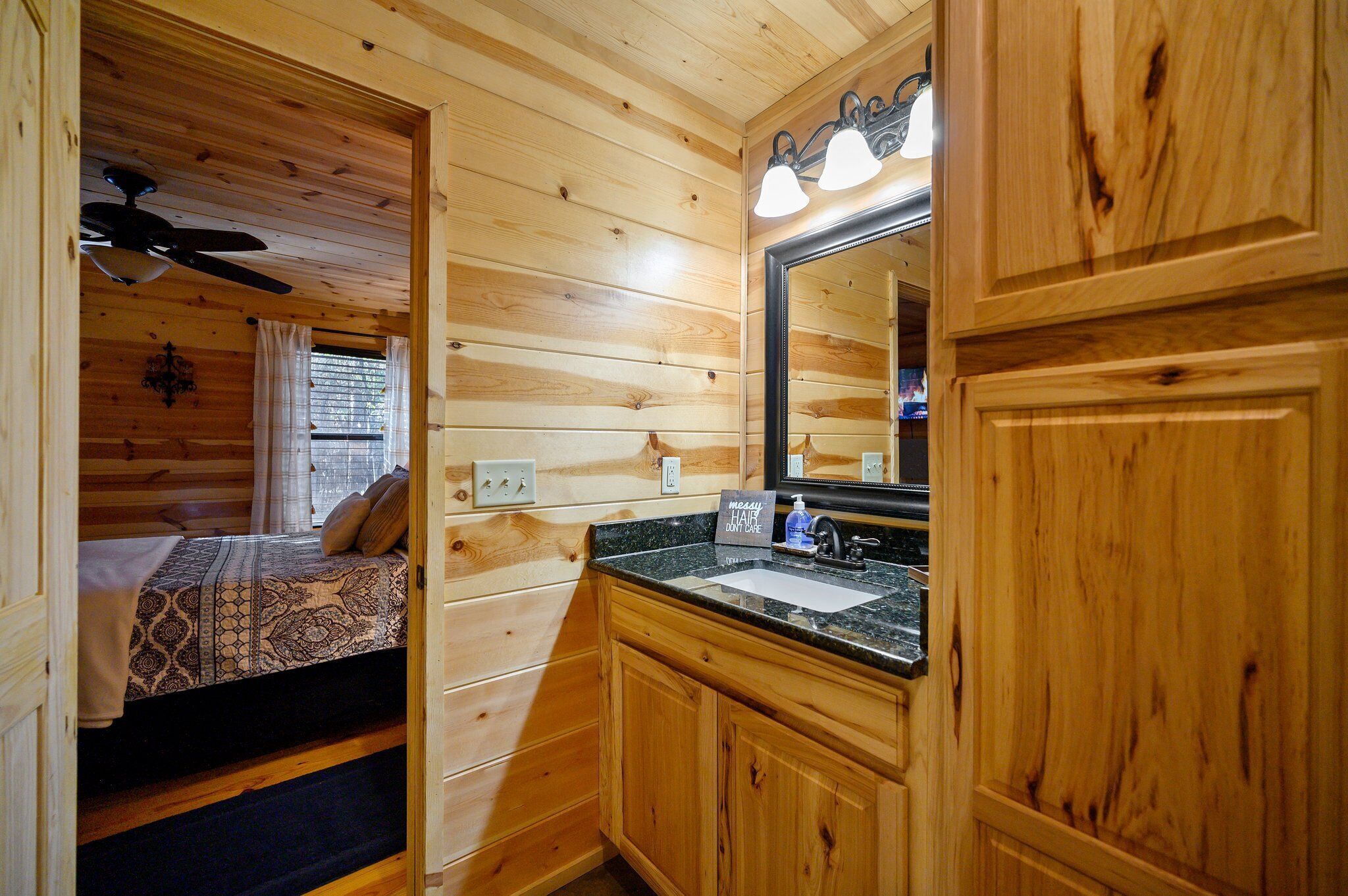 A wooden cabin interior with a bed, a bathroom with a sink and mirror, and a ceiling fan.