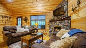 A cozy log cabin interior with a stone fireplace, leather sofas, and a wooden ceiling.