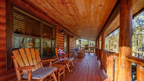 A wooden deck with log walls and large windows, featuring outdoor seating and a view of trees.