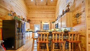 A wooden cabin interior with a kitchen area featuring a refrigerator, stove, and bar stools.