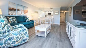 A modern living room with a teal sofa, a white coffee table, and a dining area with white chairs.