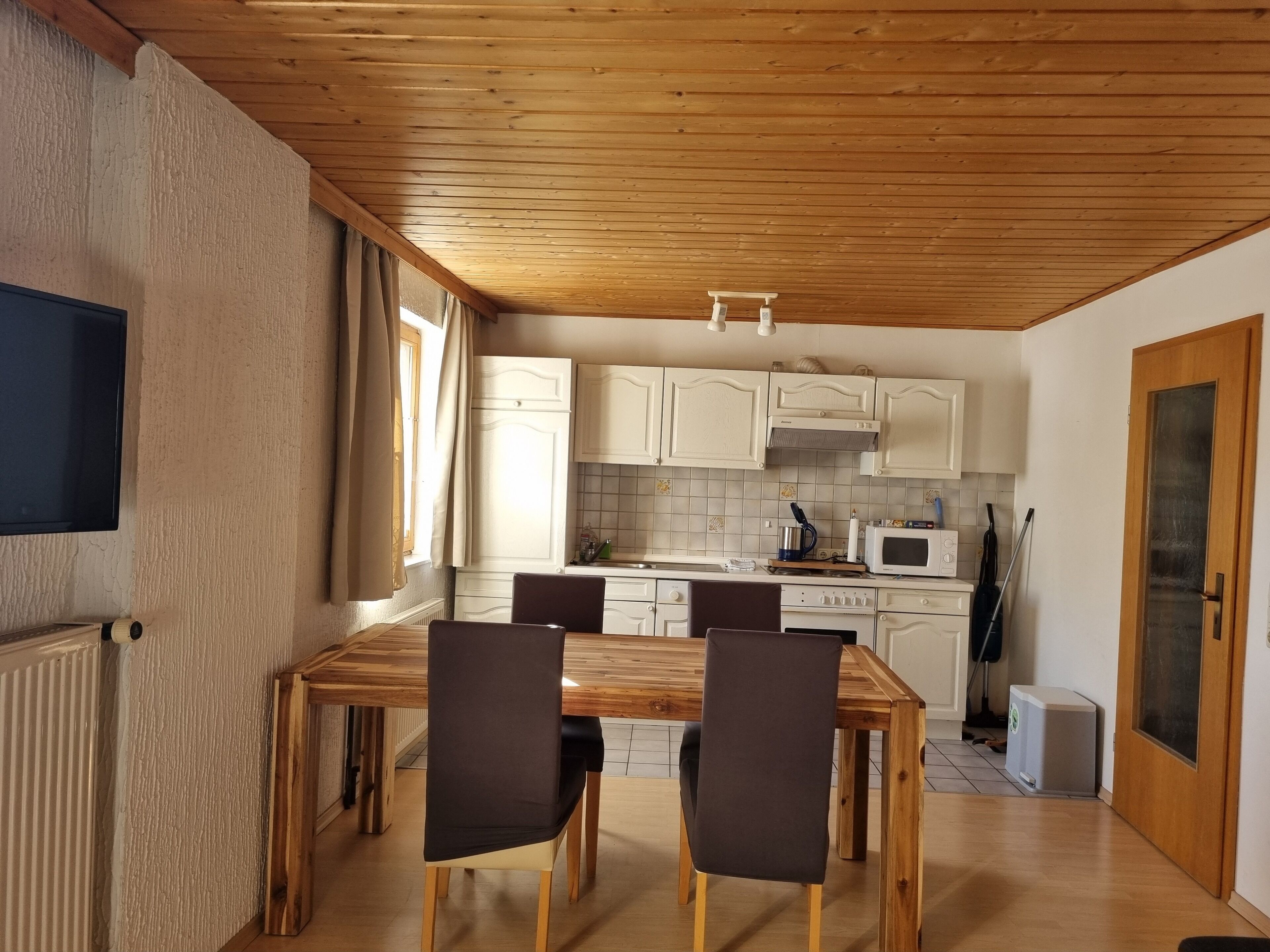 A dining area with a wooden table and chairs, a kitchen with white cabinets and a microwave, and a wooden ceiling.