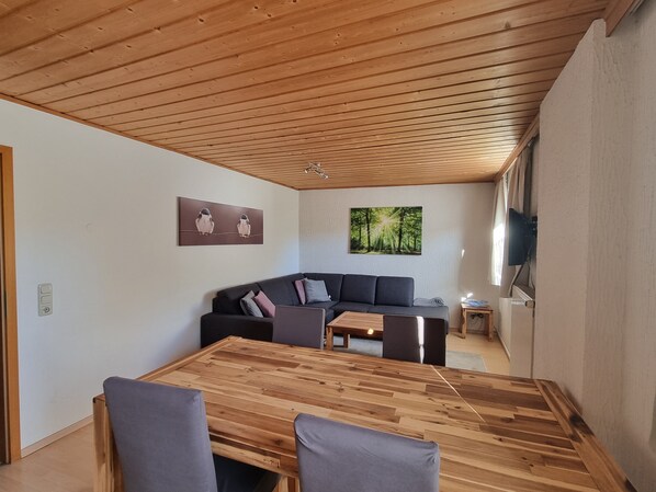 Ett modernt vardagsrum med ett träpanelat tak, en mörkgrå soffa, ett träbord och två tavlor på väggarna.