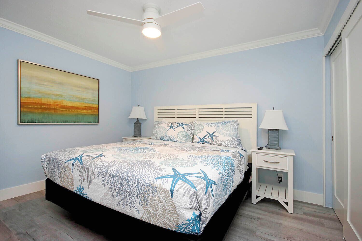 Une chambre à coucher avec un lit, des tables de chevet, un ventilateur de plafond et un tableau accroché au mur.