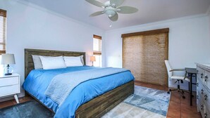 A bedroom with a wooden bed, a desk with a chair, a ceiling fan, and a window with blinds.
