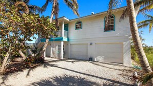 A two-story house with a front garage, surrounded by palm trees and a gravel driveway.