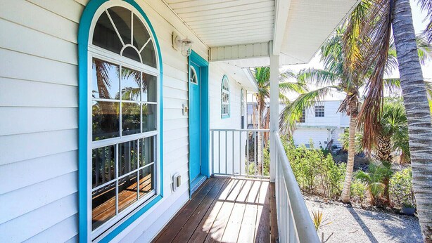 A porch with a wooden deck, a white building with blue trim, and a view of palm trees and a beach.