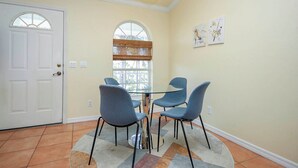 A dining area with a glass table and four chairs, a white door, a window with blinds, and two wall-mounted artworks.