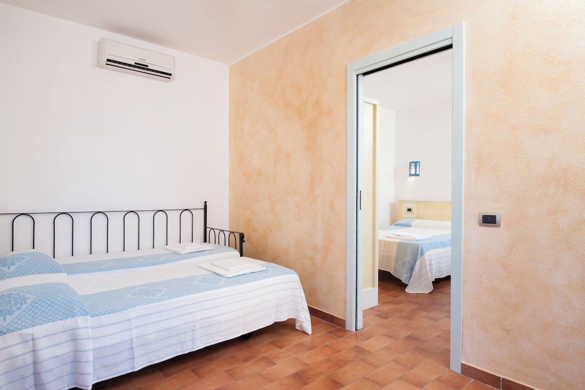 Une chambre à coucher comprenant un lit, un appareil de climatisation, une porte donnant sur une autre pièce et un sol carrelé.