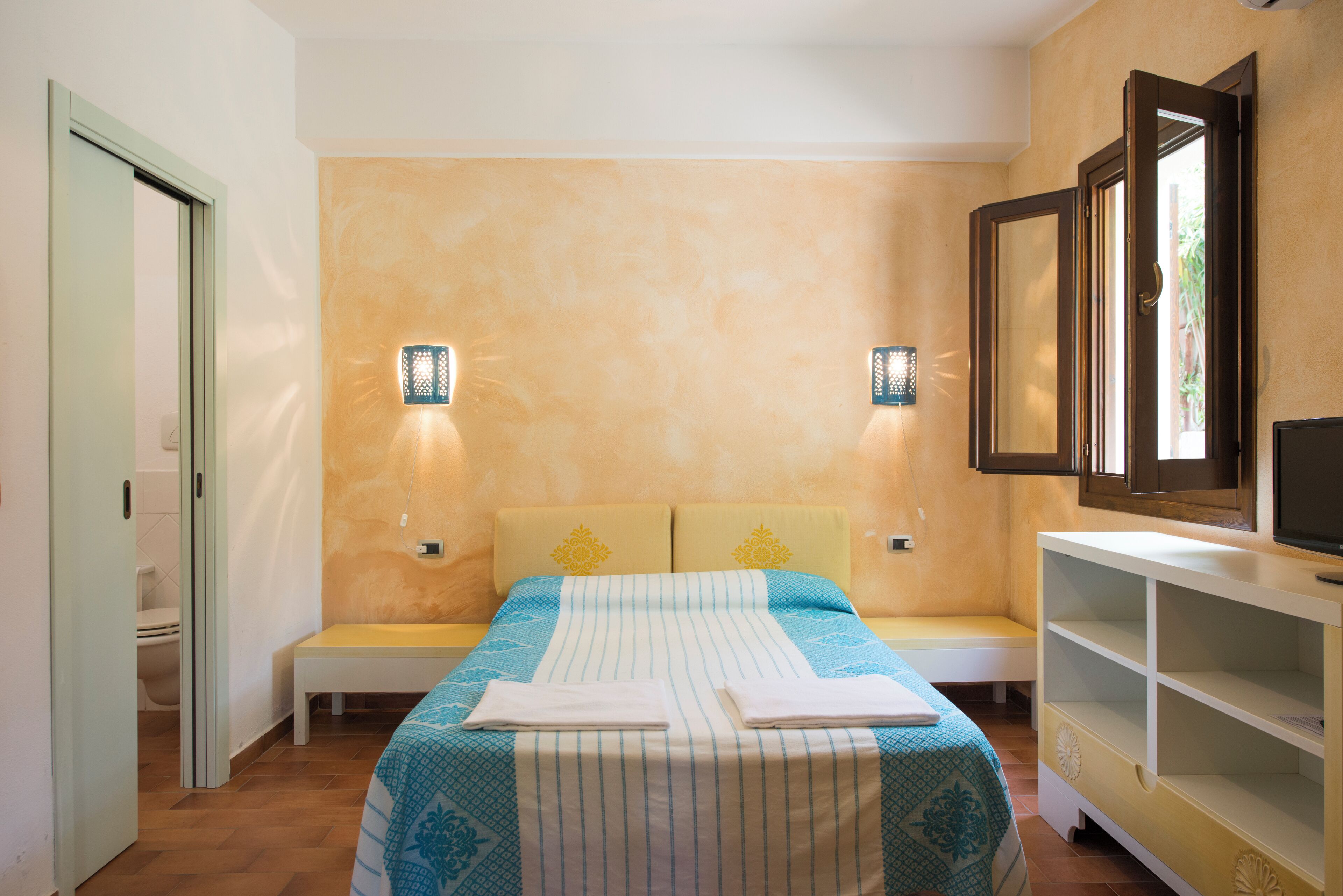 A bedroom with a bed, bedside tables, a bench, a TV, and wall-mounted lights.