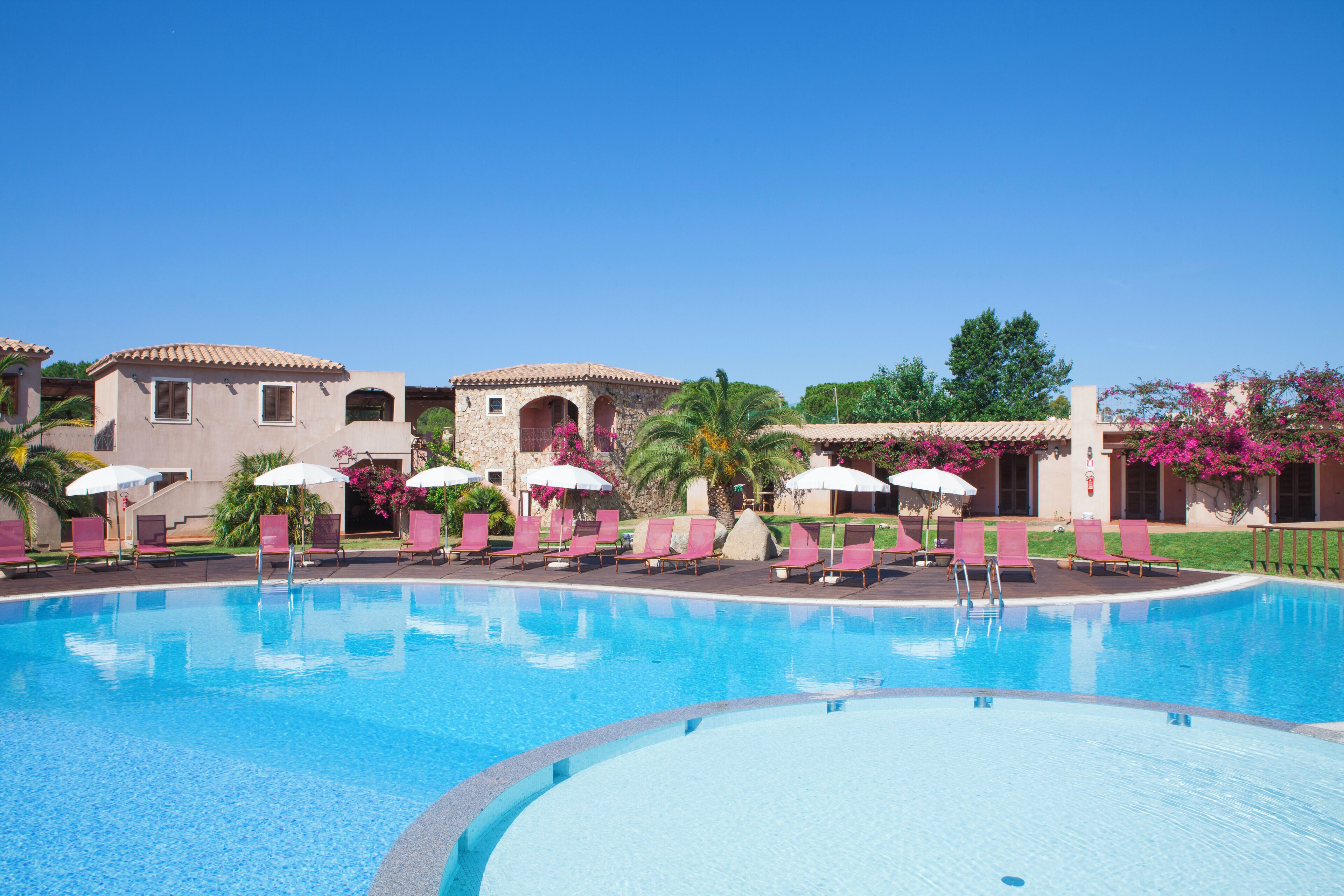 A swimming pool with pink lounge chairs and umbrellas, surrounded by buildings with arched windows and a clear blue sky.