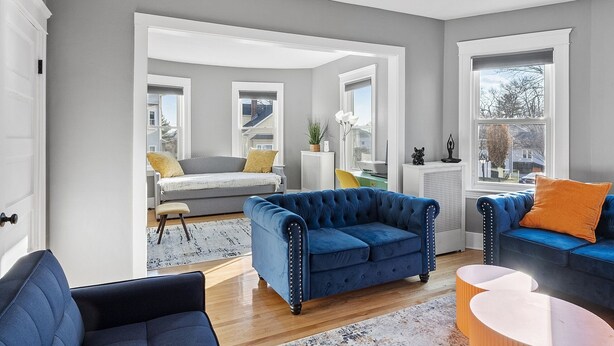 A modern living room with a blue sofa, a yellow chair, and a blue armchair. There is a white door and a window with a view of the outside.