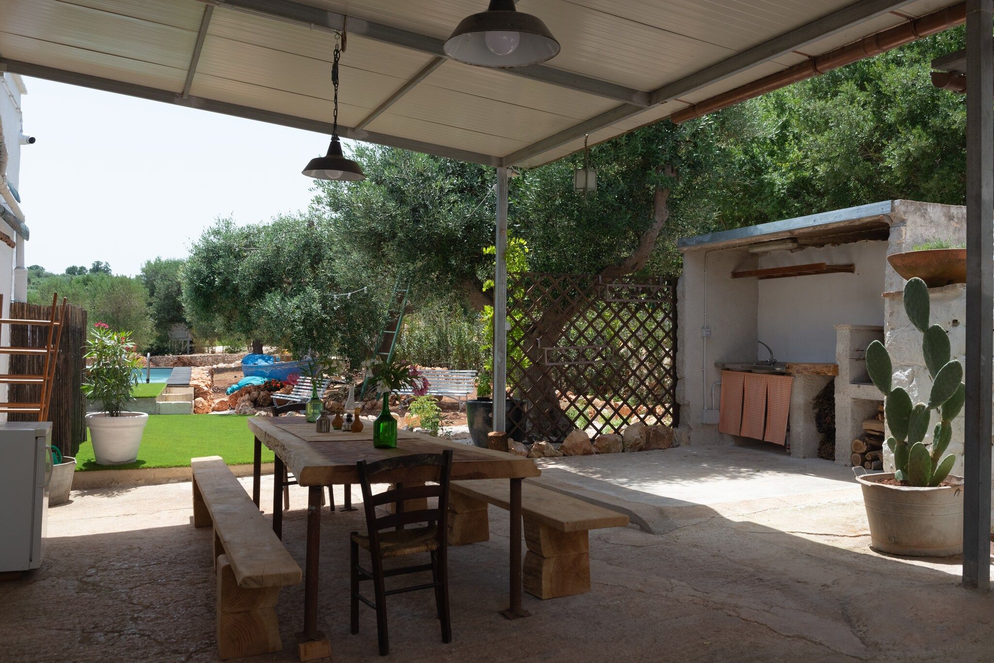 An outdoor patio with a table and benches, surrounded by a garden and a fenced area.