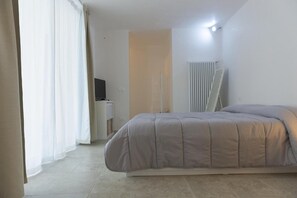A bedroom with a bed, a TV on a stand, a radiator, and a window with curtains.