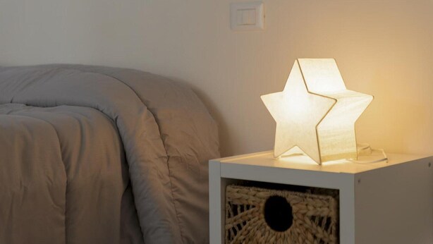 En seng med en grå dyne, et hvidt natbord med en stjerneformet lampe og en flettet kurv på natbordet.