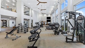 Un gimnasio espacioso con varios equipos de ejercicio, amplias ventanas y un techo alto con ventiladores.