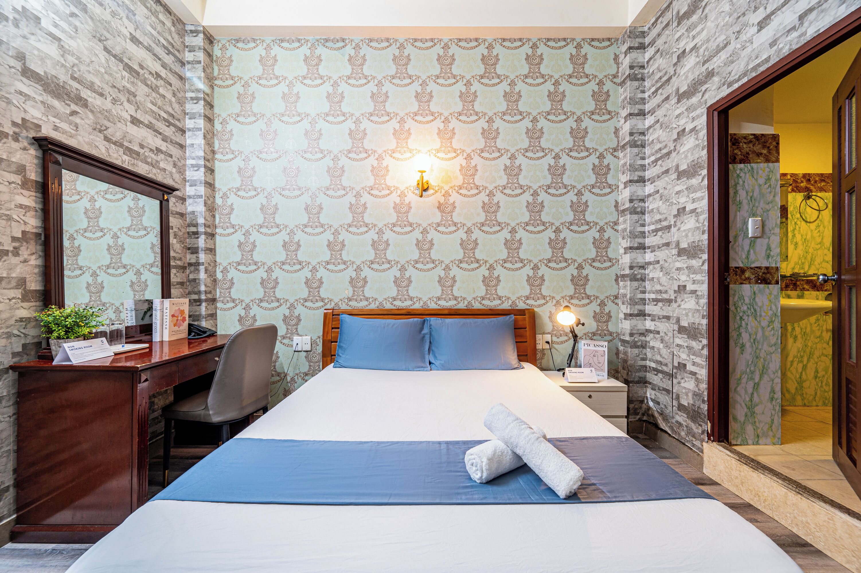 A hotel room with a large bed, a desk with a chair, a desk lamp, a nightstand, a mirror, and a bathroom visible through an open door.