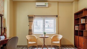 Una habitación con dos sillones de mimbre, una pequeña mesa redonda con una planta en maceta, una estantería de madera y una ventana con cortinas.