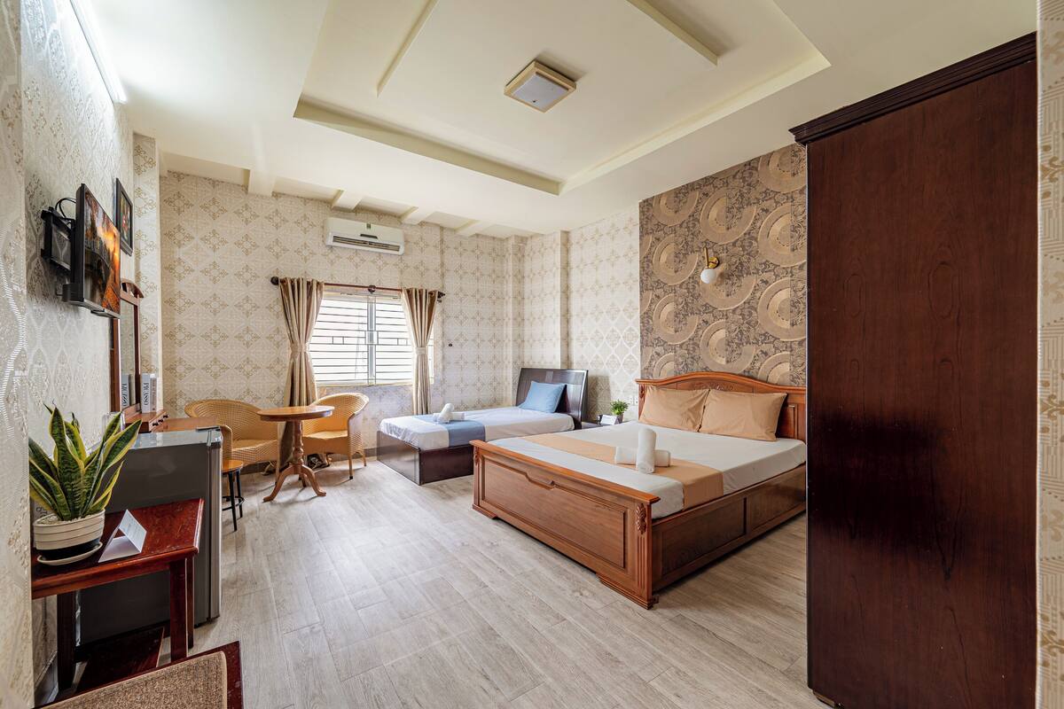 A spacious room with a large bed, a sofa, a small table, and a desk. There is a potted plant on the desk and a window with curtains.