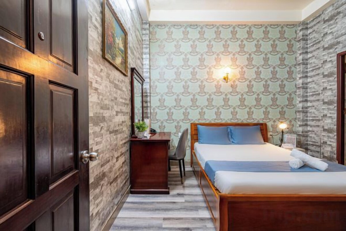 A bedroom with a bed, a desk, and a chair. There is a door on the left and a wall with a patterned wallpaper.
