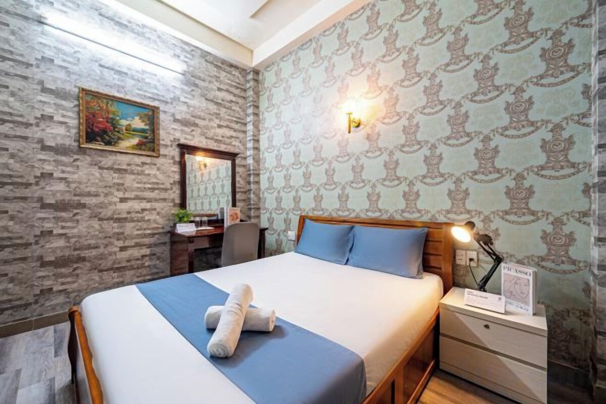 A hotel room with a bed, a nightstand, a desk, and a wall-mounted lamp.