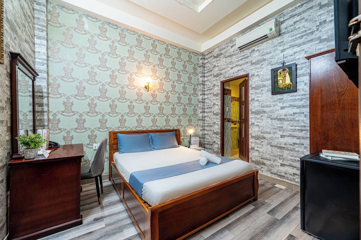 A bedroom with a wooden bed, a desk, a chair, a mirror, and a wall-mounted light fixture.