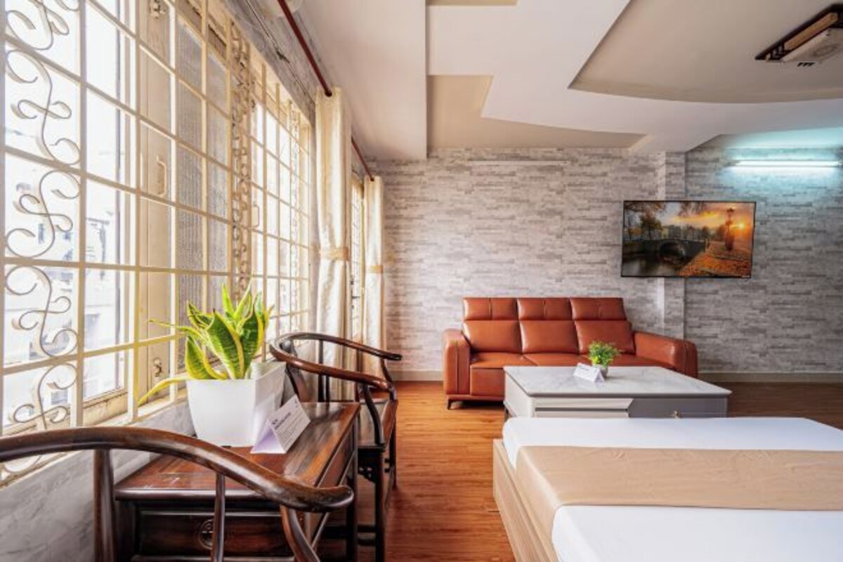 A modern hotel room with a wooden desk, a brown leather sofa, a bed, and a large window with decorative glass.