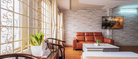 A modern hotel room with a wooden desk, a brown leather sofa, a bed, and a large window with decorative curtains.