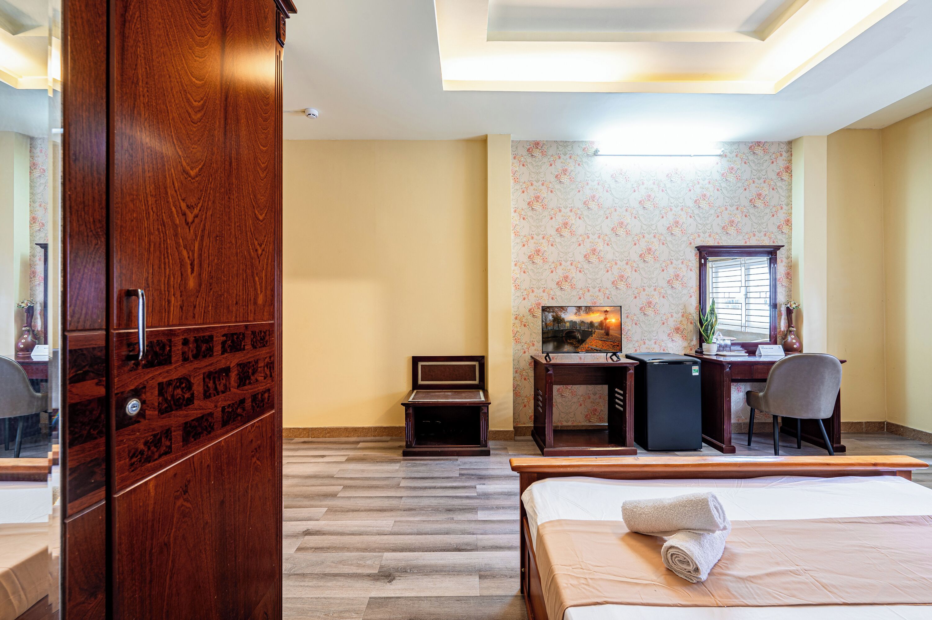 Habitación de hotel con un armario de madera, una cama con una toalla enrollada, un escritorio con una computadora, una silla y una pequeña mesa con una planta.