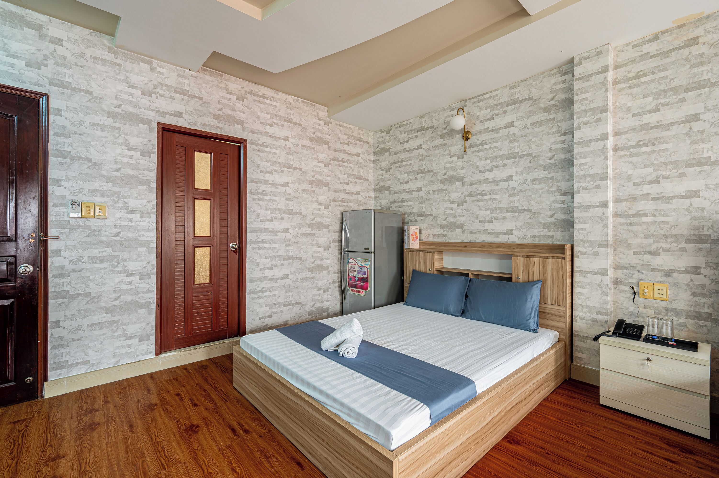 A hotel room with a wooden bed, a nightstand, a refrigerator, and a door with a glass panel.