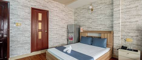 A hotel room with a wooden bed, a nightstand, a refrigerator, and a door with a glass panel.