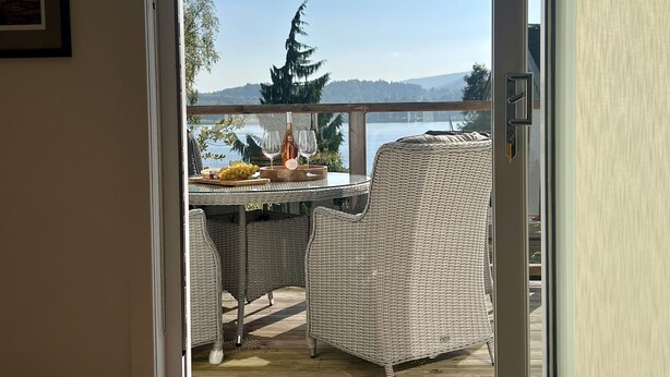 A balcony with wicker furniture, a table with fruit and wine glasses, and a view of a lake and mountains.