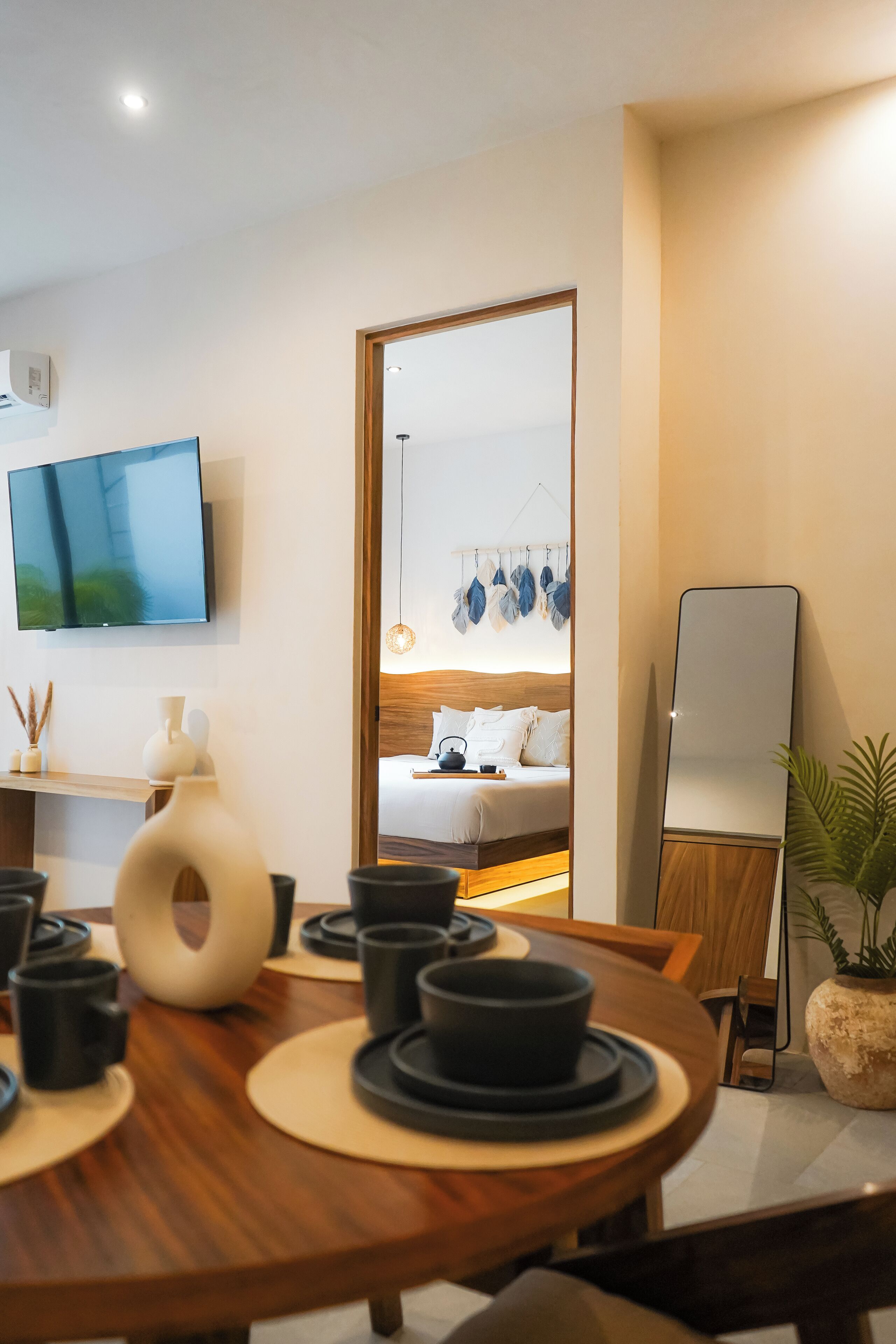 Habitació d'hotel moderna amb taula de dinar de fusta, televisió de pantalla plana, un gran mirall i un llit amb capçalera de fusta.