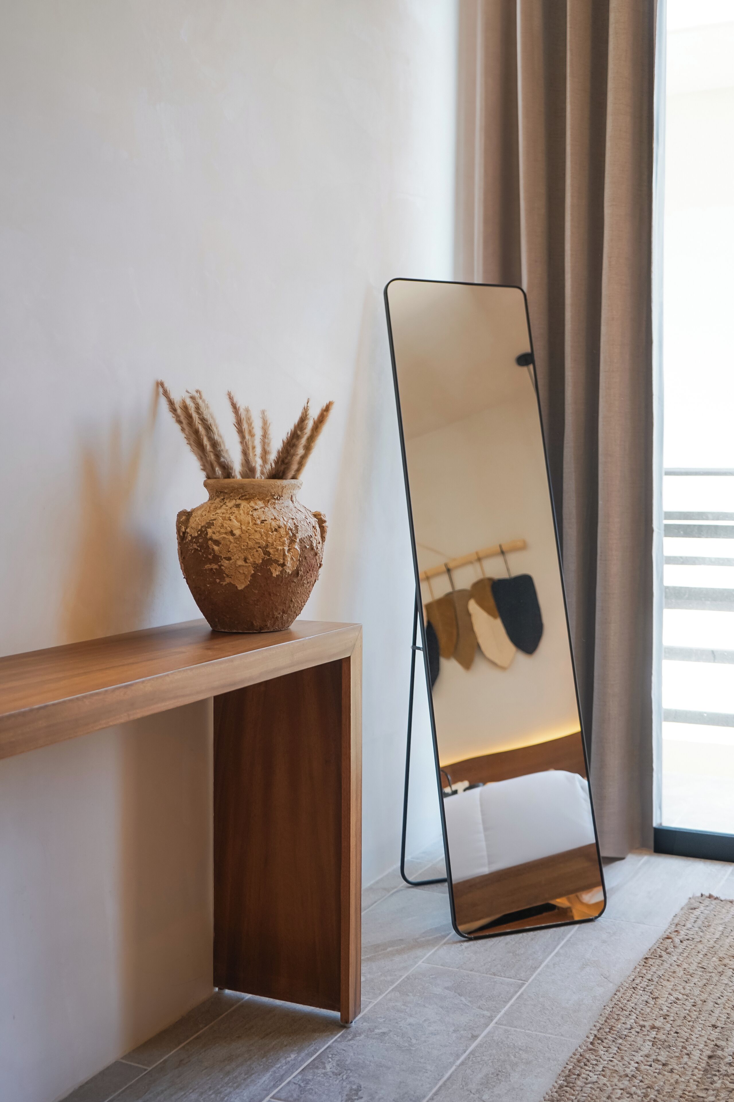 A wooden console table with a textured vase containing dried plants, a standing mirror, and a light-colored rug on a tiled floor.
