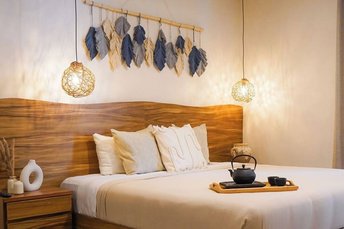 A modern bedroom with a wooden headboard, a large bed, bedside tables, and a decorative leaf wall hanging.