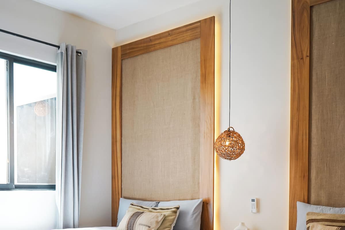 A hotel room with two beds, a large window with curtains, a wooden headboard, a woven light fixture, and a name tag on the bed.