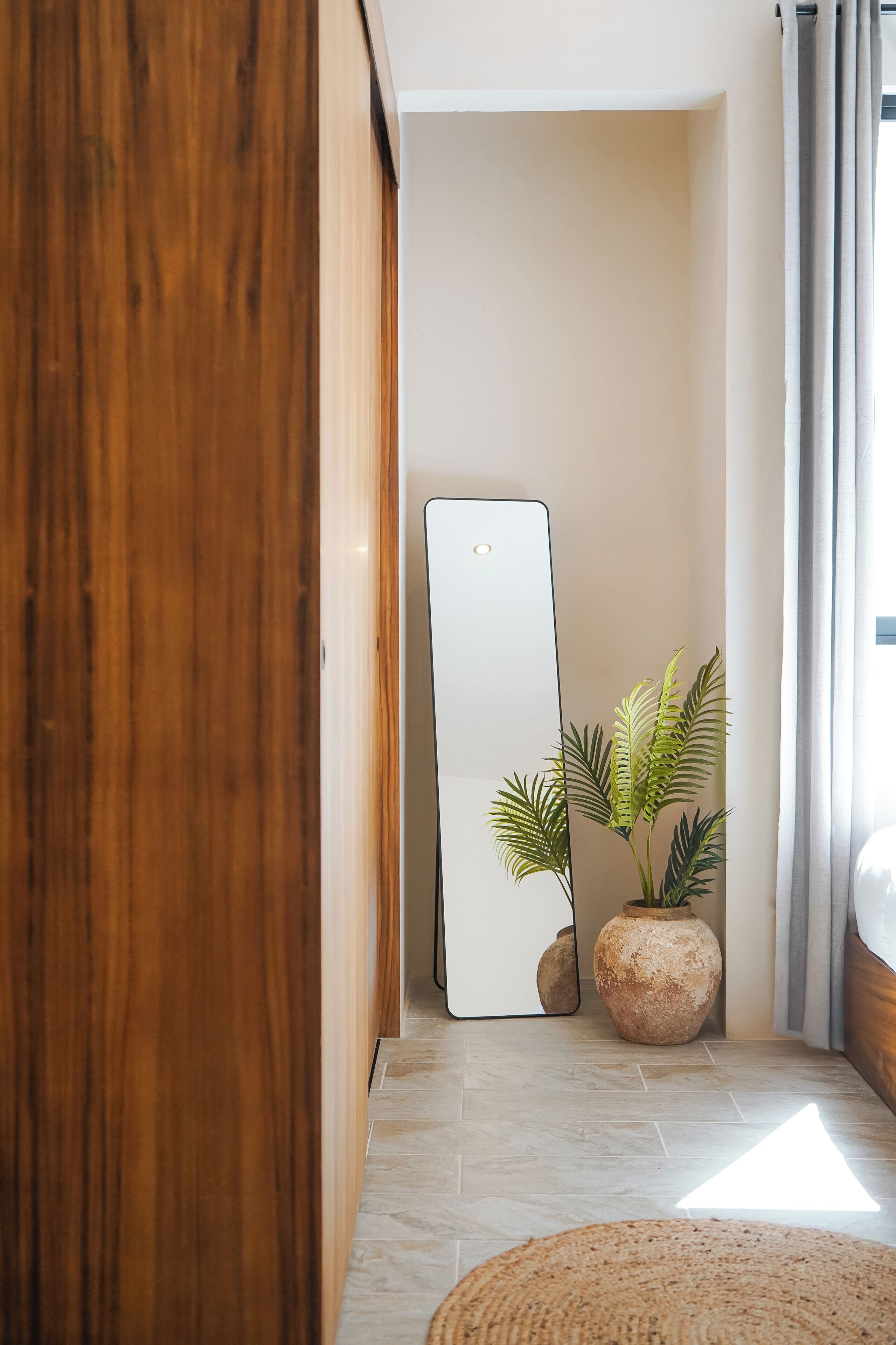 Un corridoio con una porta in legno, uno specchio con cornice bianca, una pianta in vaso e un tappeto rotondo intrecciato.