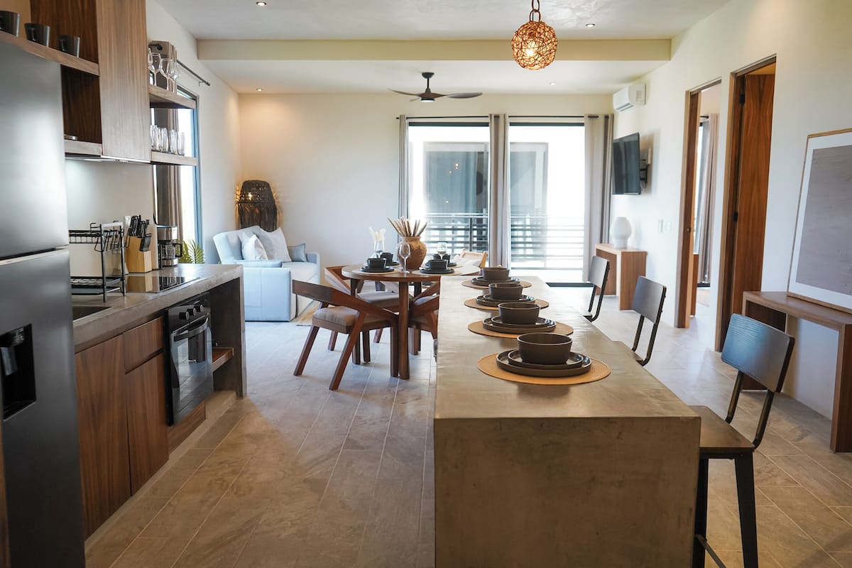 A modern kitchen with a central island, dining area with chairs, and a large window providing natural light.