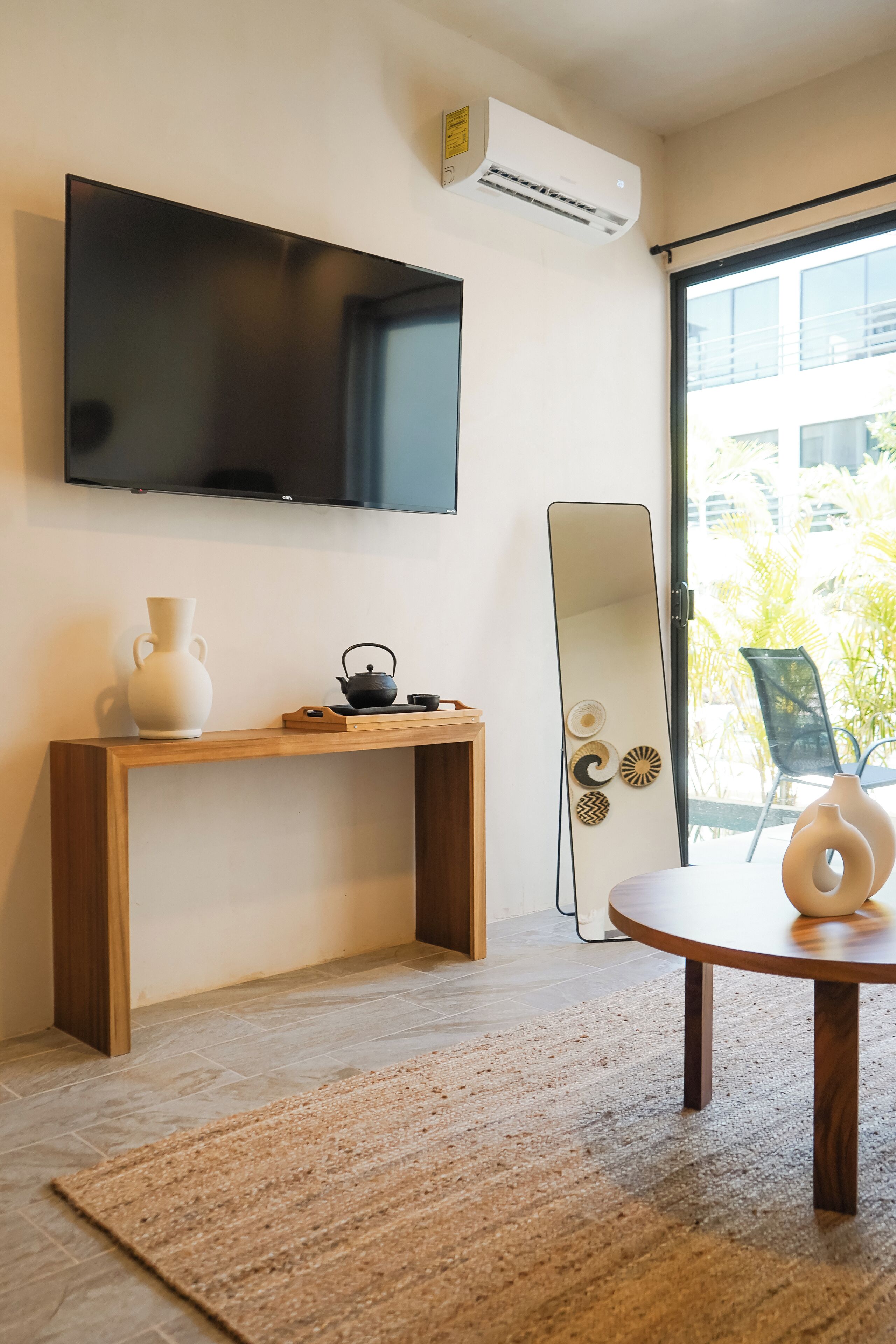 A modern living room with a flat-screen TV, a wooden console table with decorative items, a round table with a vase, and a large mirror.