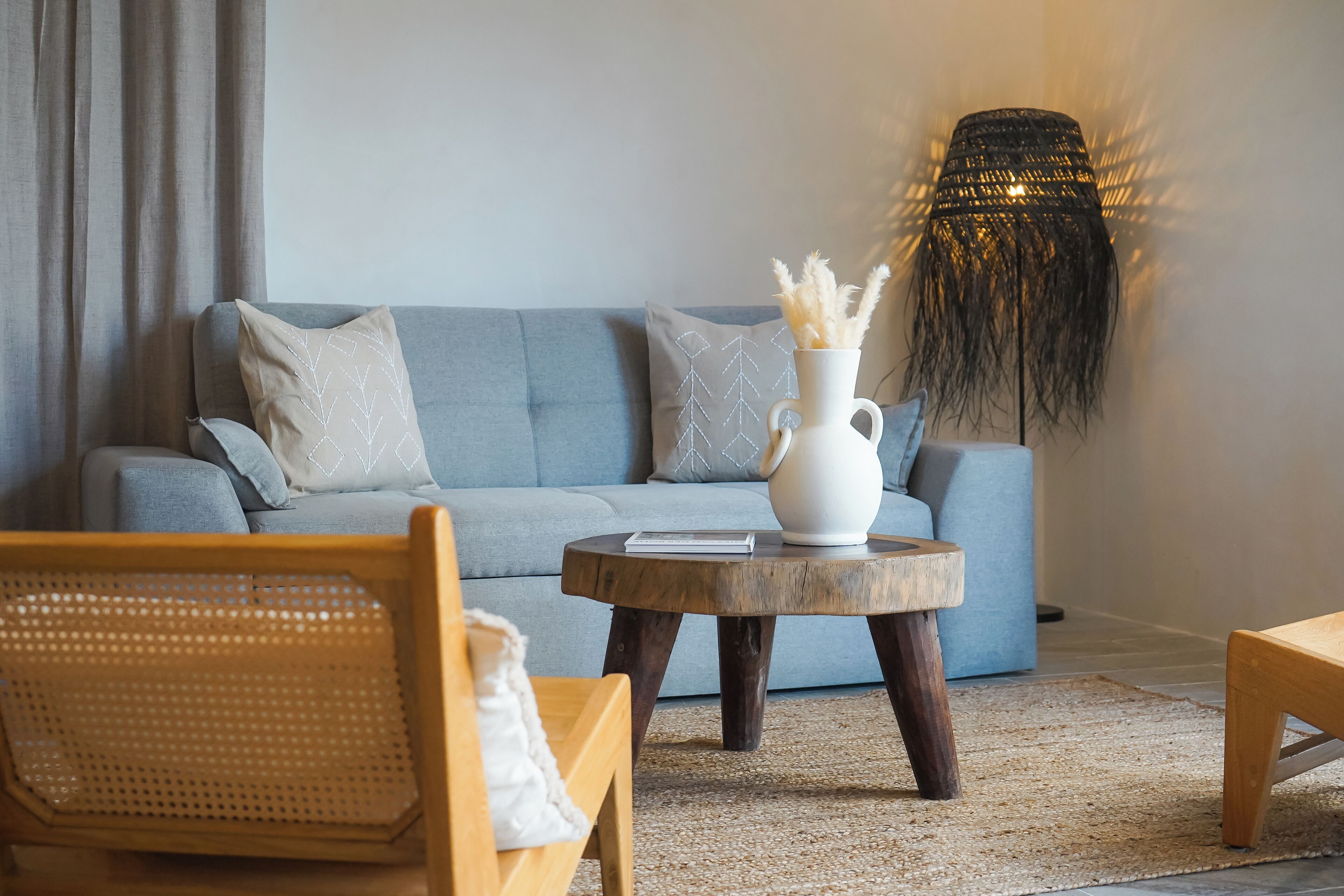 Una sala de estar con un sofá gris, una mesa de centro redonda de madera, una silla de mimbre y una lámpara de pie.