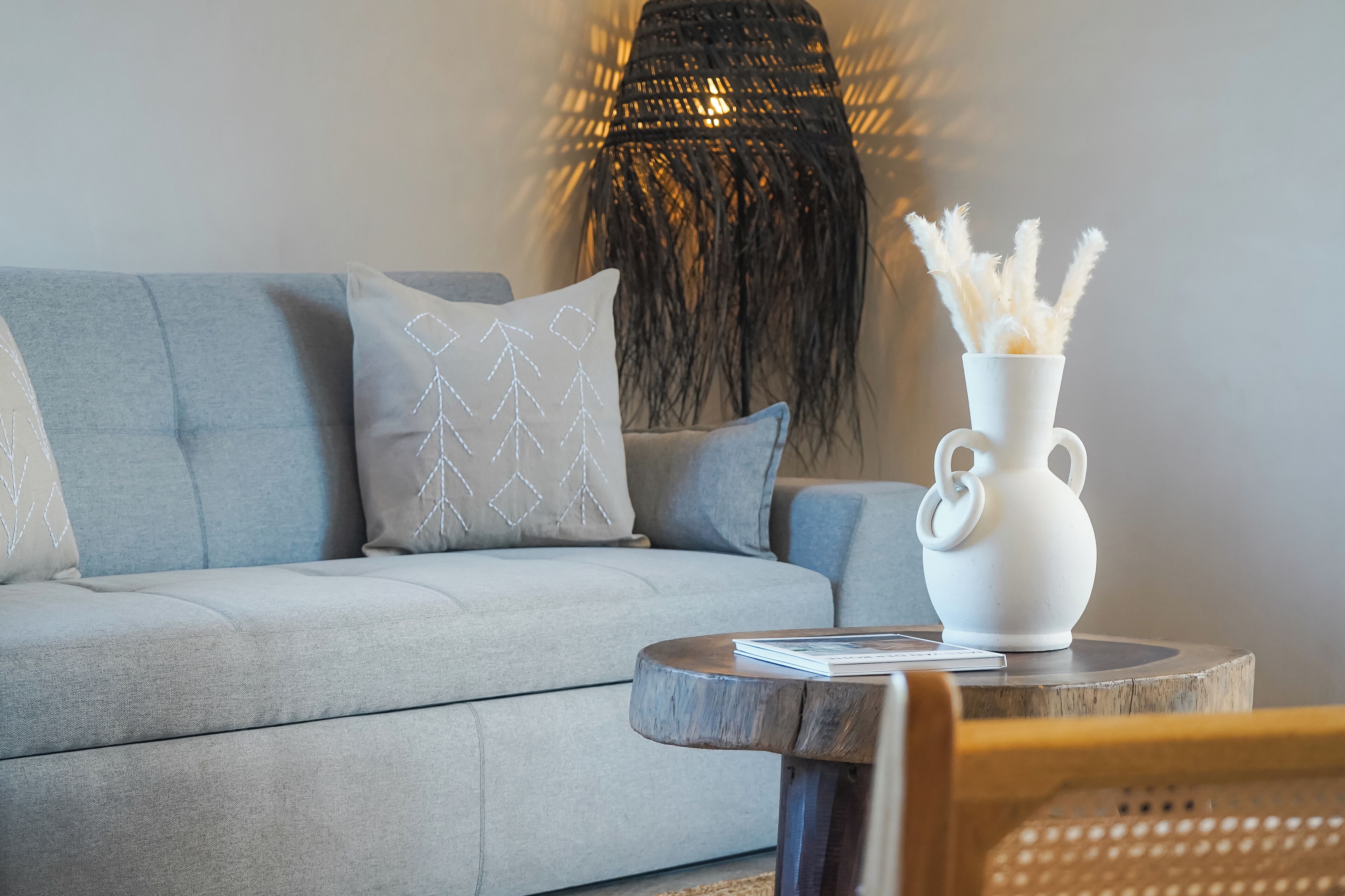 Un salon moderne avec un canapé gris, des coussins à motifs, un vase blanc contenant des plumes d’herbe des pampas et une table basse en bois.