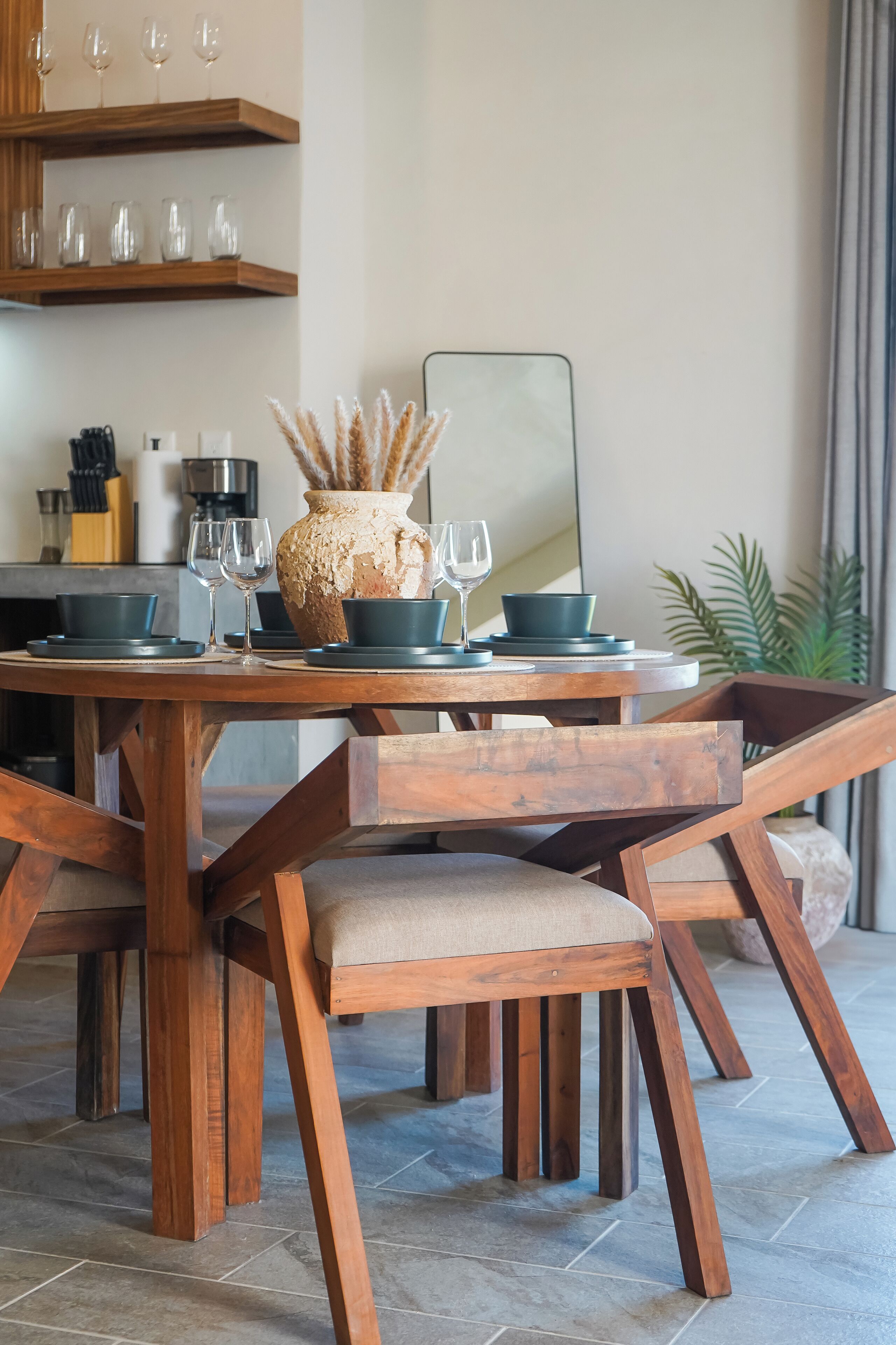 Una mesa de madera con sillas a juego, un florero con plantas secas y un espejo sobre la mesa.