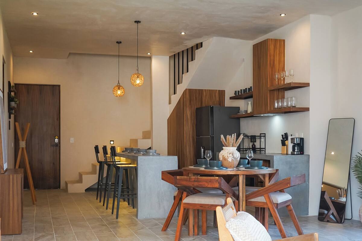 A modern kitchen with wooden cabinets, a dining area with a round table and chairs, and a staircase leading to an upper level.